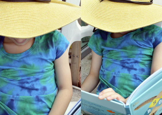 child wearing handmade tie-dye tee and reading a book with a floppy sunhat on.