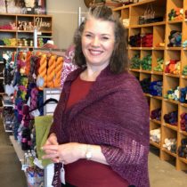 Tara Roberts, owner of Stranded by the Sea standing in her shop with a smile wearing Falling Petals Shawl knit with Sweet Georgia CashLuxe in a rich berry color.