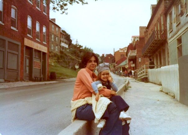 Mother with black hair and peach shirt sitting on the curb holding toddler with train conductor hat.