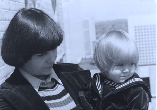 Mother with black hair and 70s style velvet jacket holding 1-year-old blonde Evin in a velvet sailor suit. As a baby, I threw up a lot so I don't know what mom mom was thinking with the velvet.