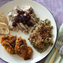 A dinner plate with roast turkey topped with cranberry sauce, some green bean mushroom casserole topped with crispy fried onions, some sweet potato casserole topped with candied pecans, and two slices of baguette. A fork and knife are beside the plate at the ready.