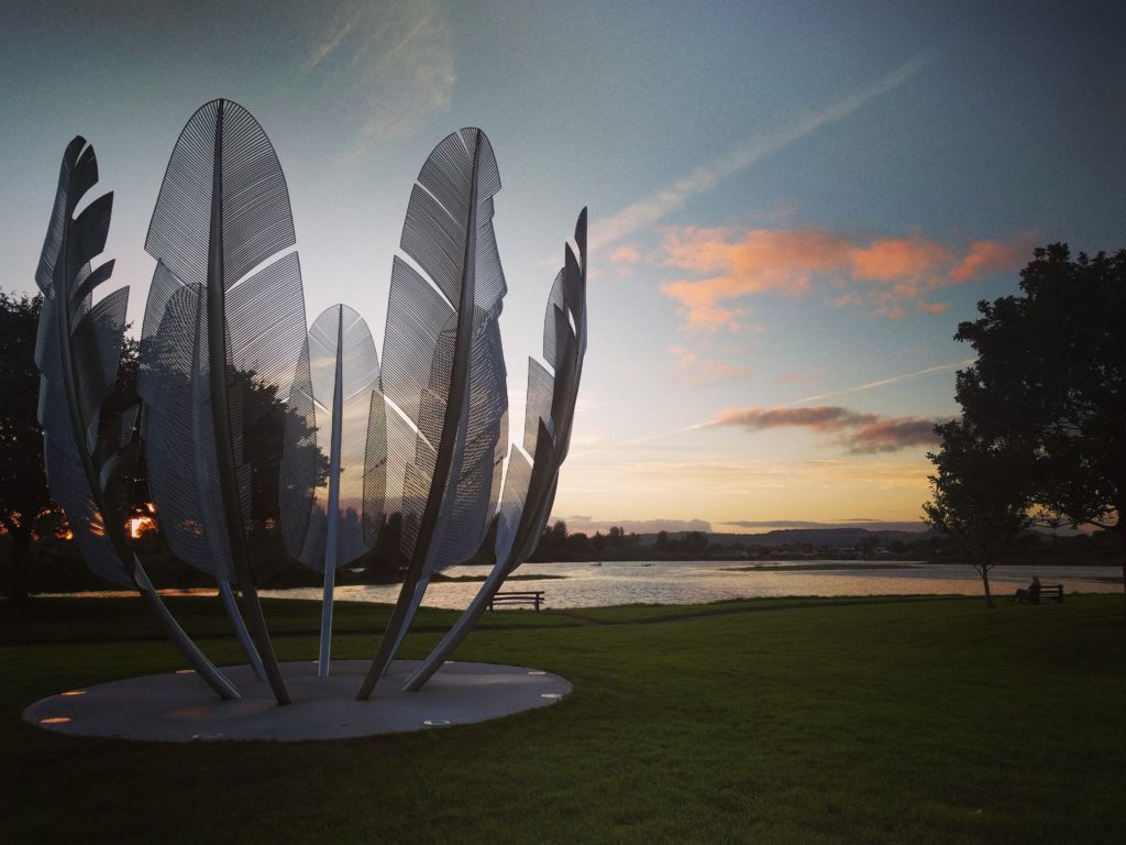 Tall metal feathers in a bowl-shape against a colourful sunset sky in Ireland