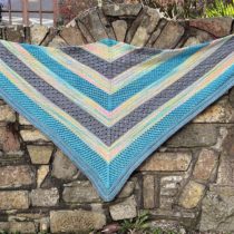 Triangle-shaped hand knit shawl against a stone wall. The bright blue, slate grey, and speckled rainbow striped on the shawl contrast boldly against the old weathered stone.
