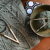 Circular wood knitting needles with sage green yarn in a bowl alongside a knit lace shawl on a wood table with a journal.