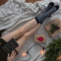 Caucasian legs outstretched on a knit blanket beside an evergreen wreath with a casual strand of lights and a couple brown paper packages with natural accents. A book in the person's lap is titled 'Your Soul is a River'