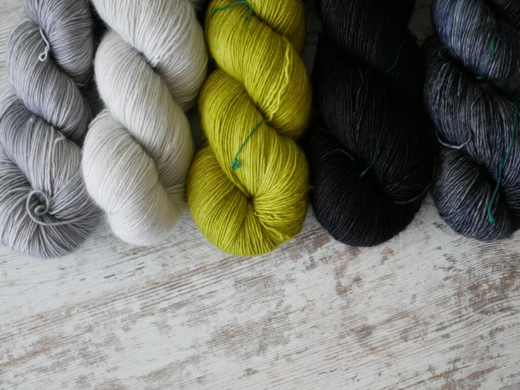 Five skeins of yarn on a white painted wood table with the wood grain showing through. The skeins are grey, white, arsenic green, black, and asphalt from left to right.