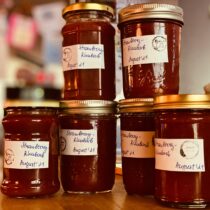Homemade jam in jars with labels and dates. This batch is Strawberry-Rhubarb and dated August '21. A small stamp on the label likely signifies the farm or maker. Photo by Max Goncharov