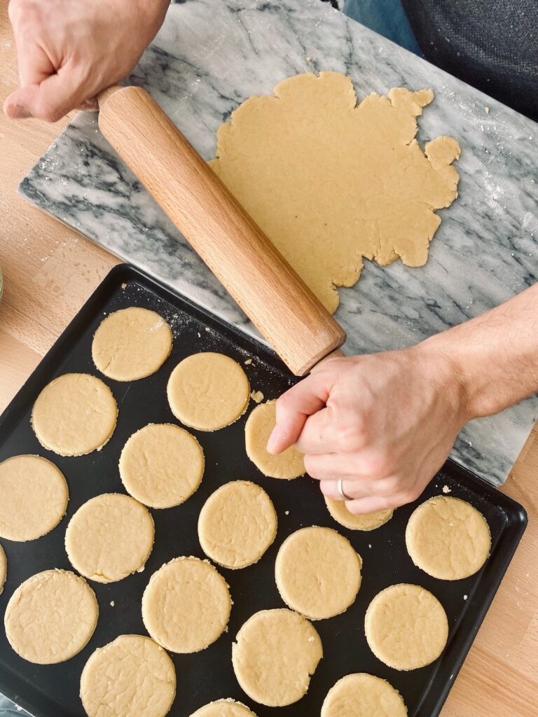 Rolling out the dough to make more shortbread cookies with a baking sheet alongside covered in round shortbread cookies about to go in the oven. 