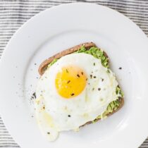 A sunny side up egg on spread avocado atop toast on a white plate.